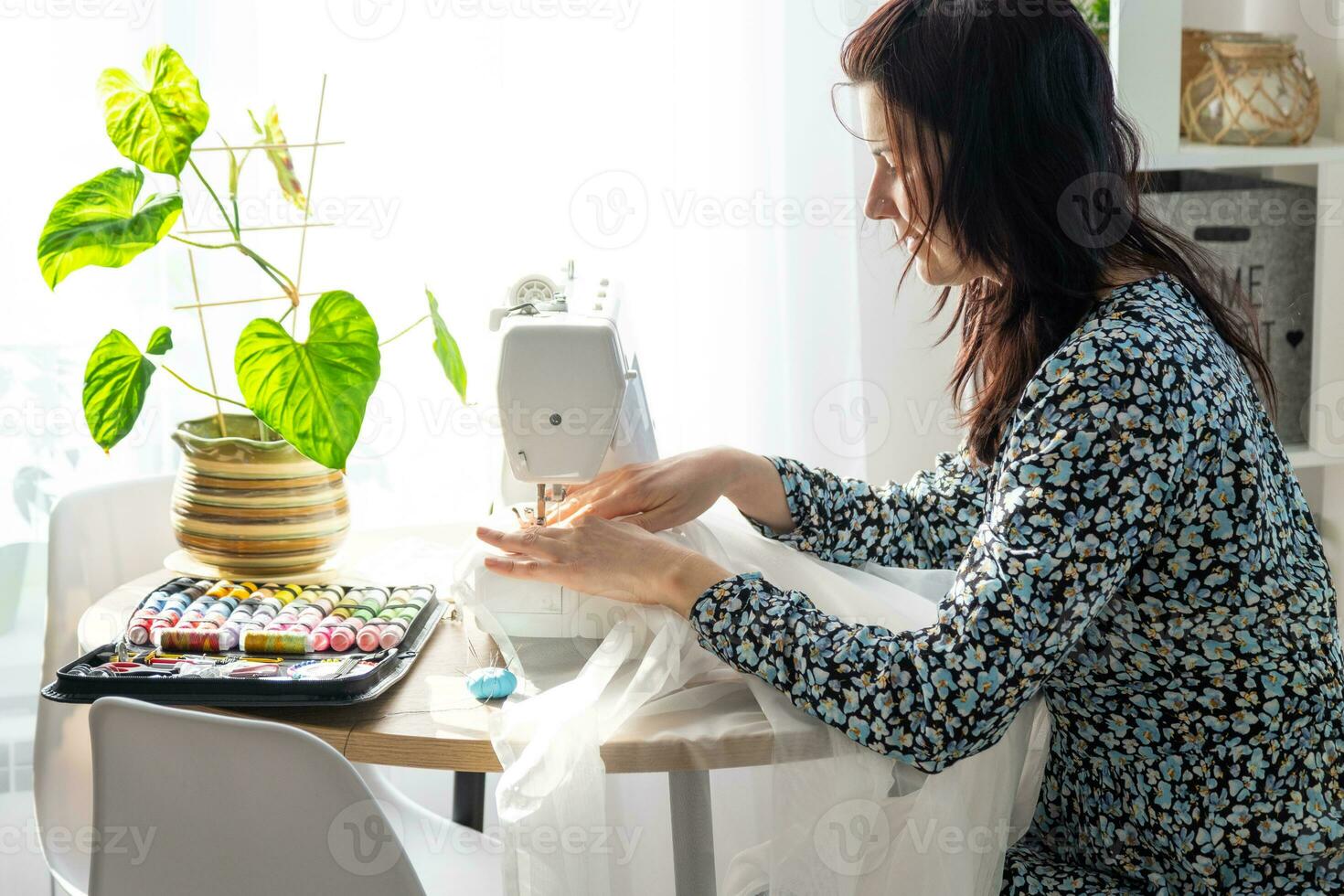 een vrouw naait tule Aan een elektrisch naaien machine in een wit modern interieur van een huis met groot ramen, huis planten. comfort in de huis, een huisvrouw hobby foto