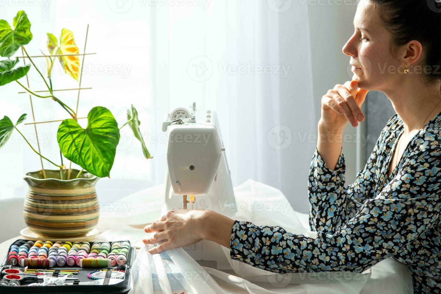 vrouw denken, dromen en naait tule Aan een elektrisch naaien machine in een wit modern interieur van een huis met groot ramen, huis planten. comfort in huis, huisvrouw hobby foto