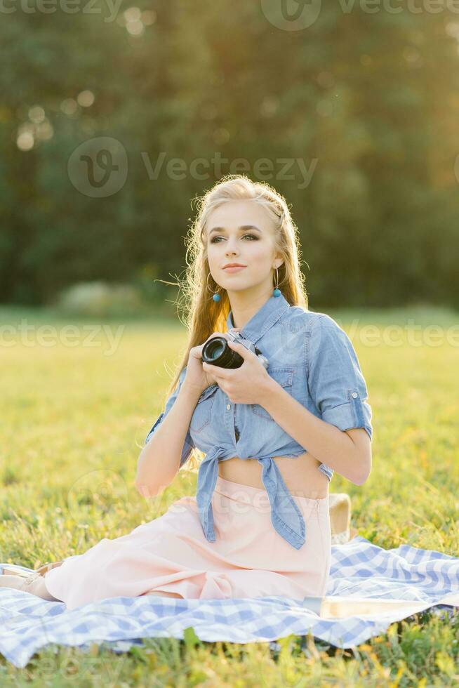 een jong mooi vrouw zit Aan een deken in natuur en houdt een camera in haar handen foto