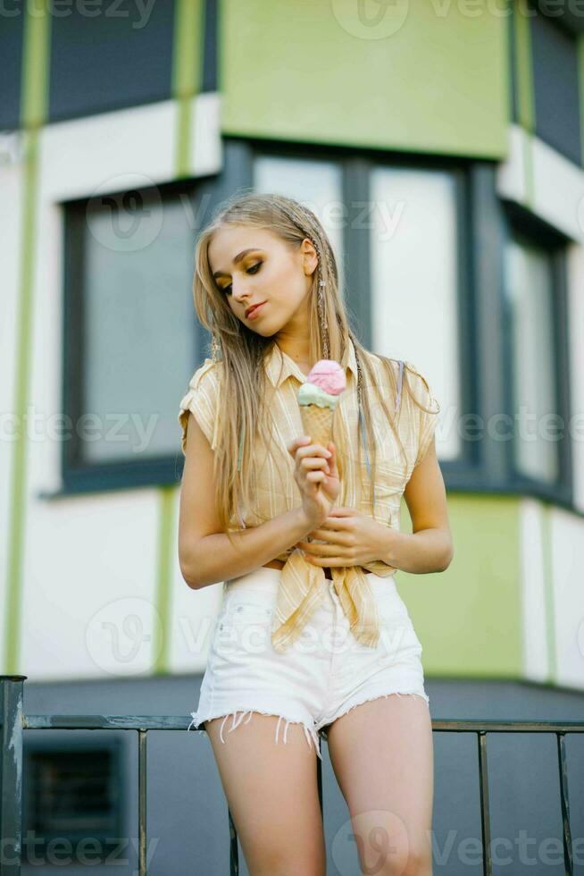 een jong mooi vrouw houdt een ijs room in een wafel ijshoorntje tegen de achtergrond van stad huizen in zomer foto