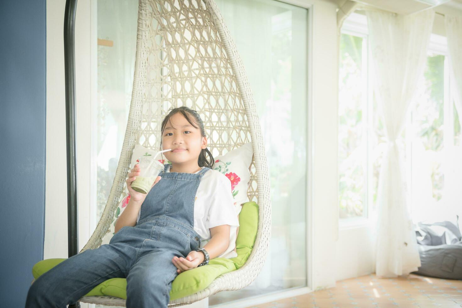 schattig meisje vervelend een denim overall is drinken ijs groen thee en kom tot rust in wit huis. foto
