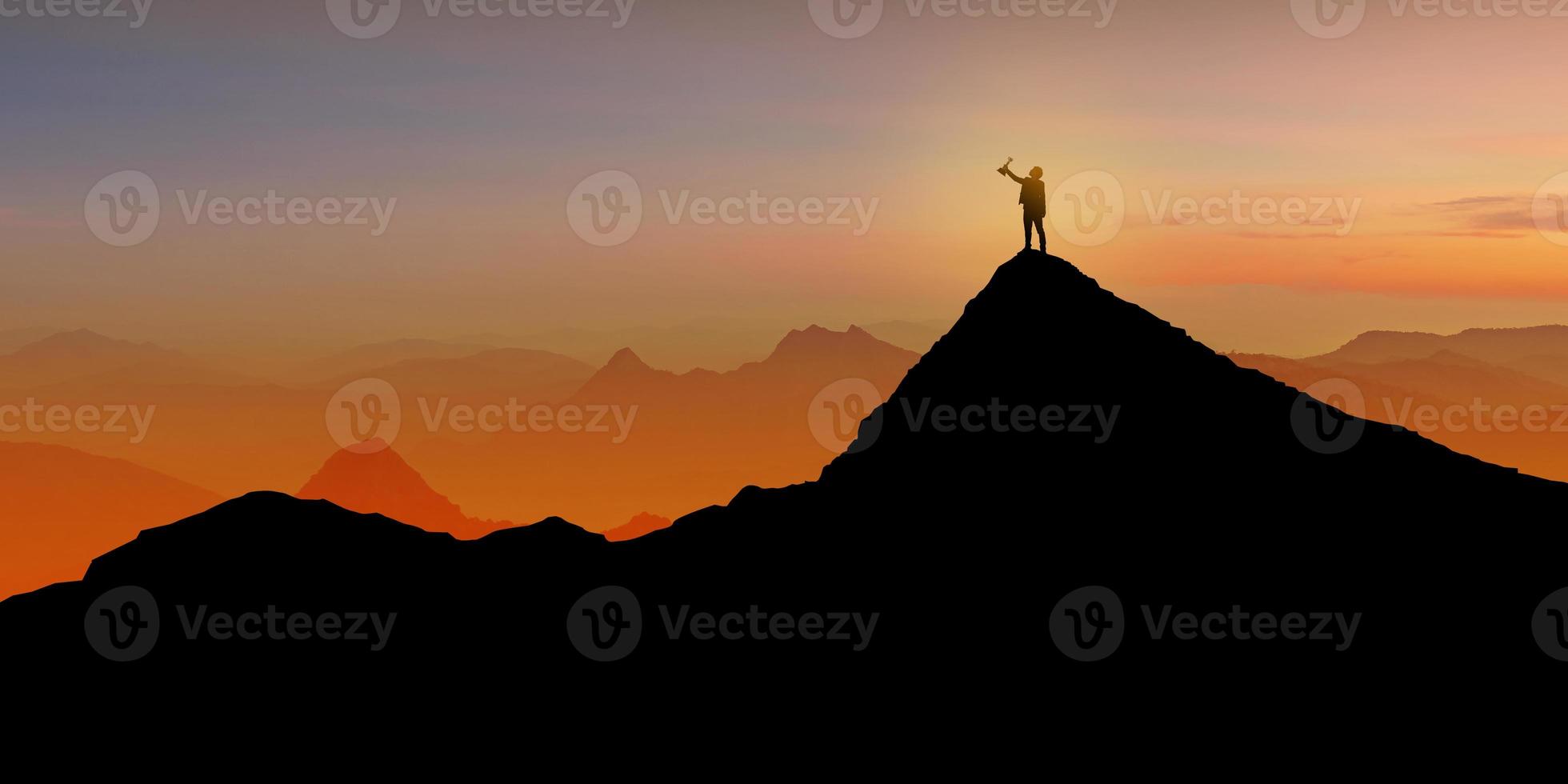 silhouet van zakenman staande op de bergtop over zonsopgang schemering achtergrond met een trofee cup omhoog te houden foto