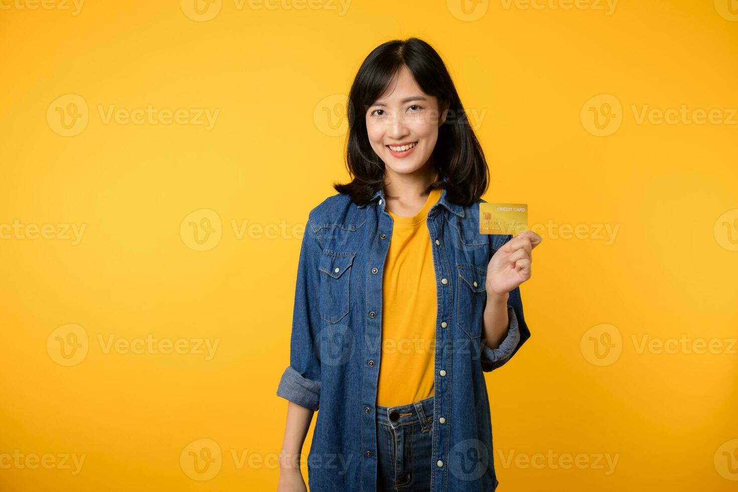 portret van jong Aziatisch vrouw vervelend geel t-shirt en denim jasje met gelukkig glimlach Holding een credit kaart geïsoleerd Aan geel achtergrond. betaling boodschappen doen online concept. foto