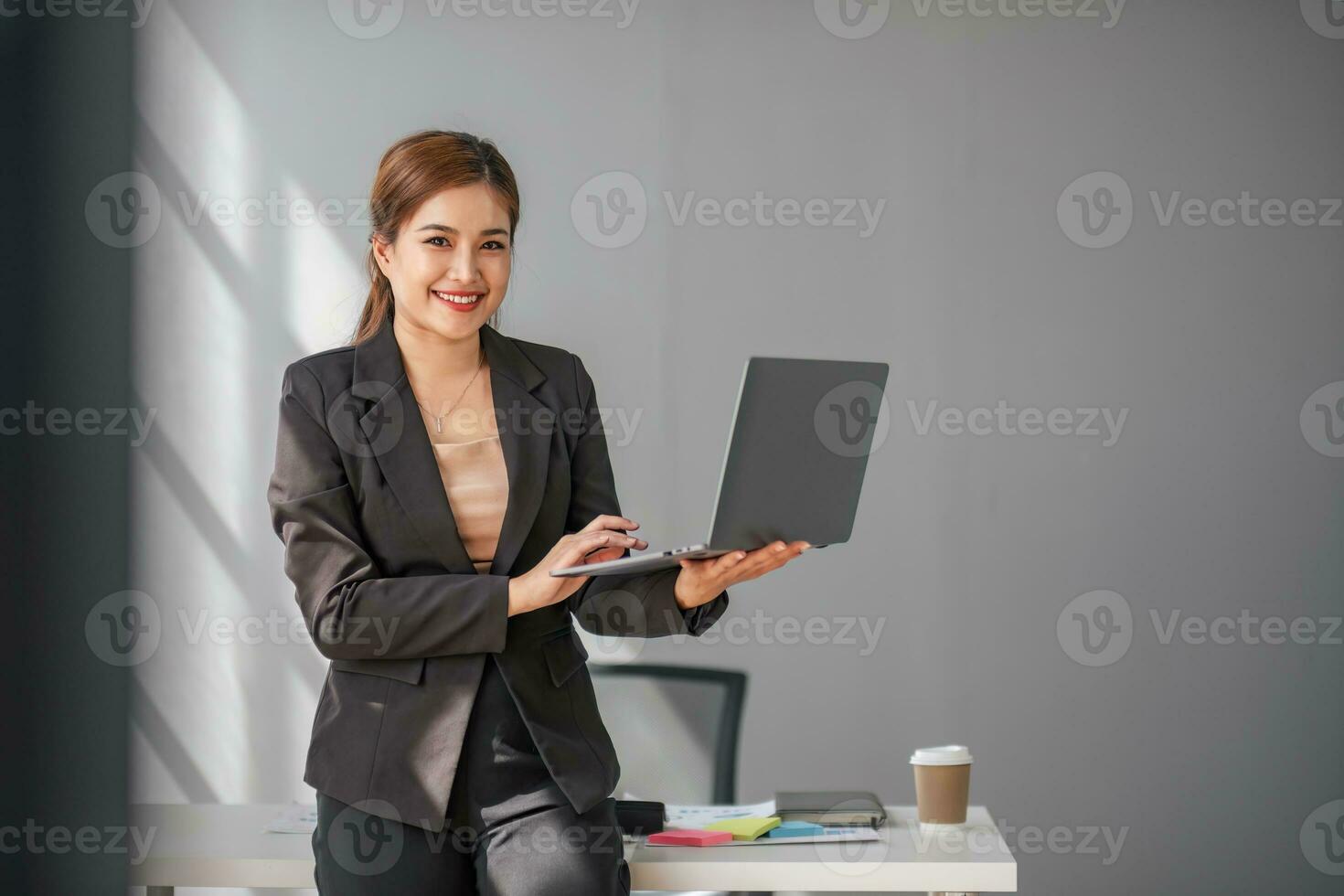 portret, professioneel en zelfverzekerd Aziatisch zakenvrouw of vrouw uitvoerend manager in formeel pak en gebruik laptop. staan, leunend Aan tafel, Holding laptop en gebruik makend van laptop computer. foto