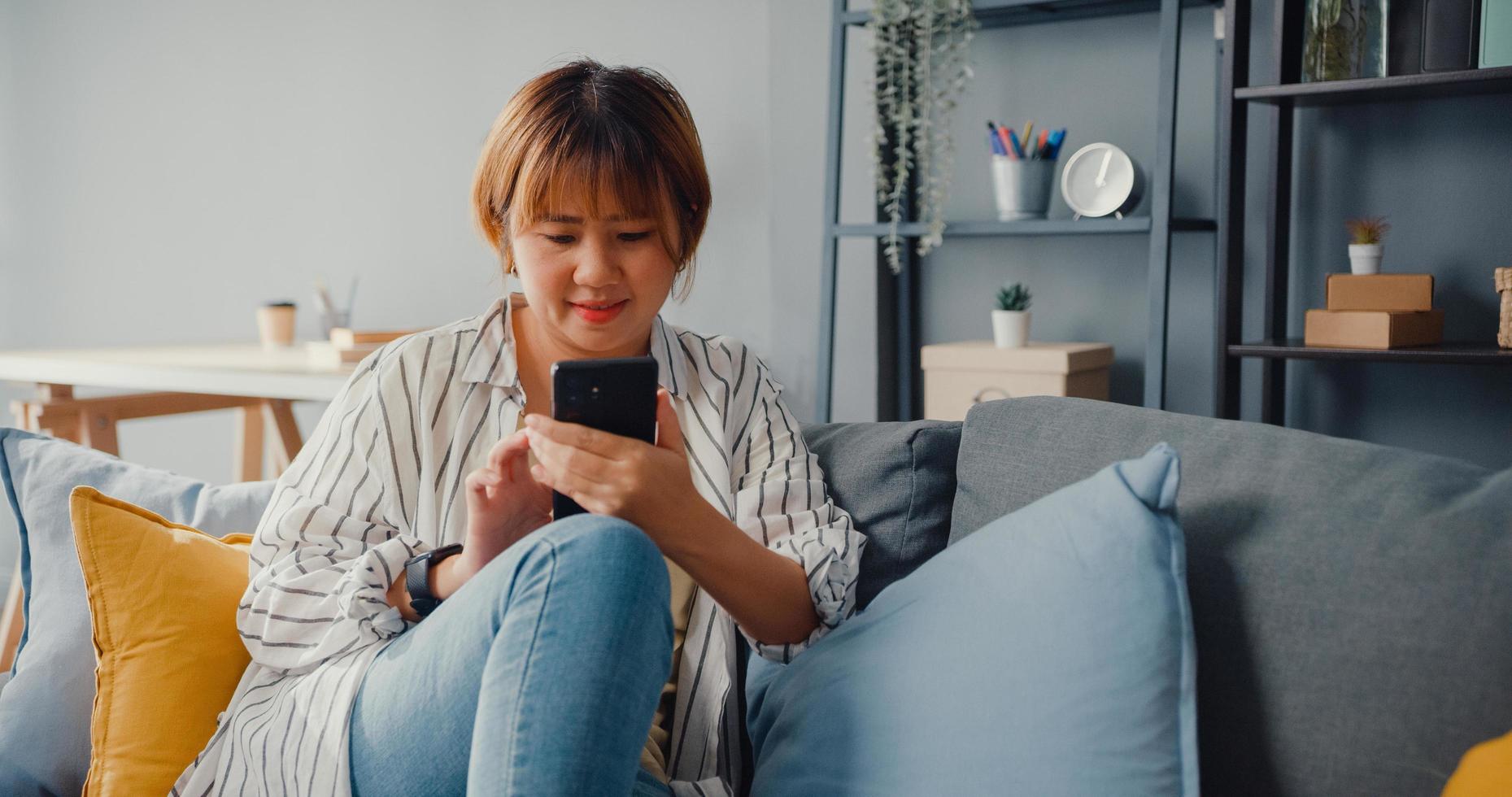 jonge Aziatische dame die smartphone-sms-bericht gebruikt of sociale media checkt op de bank in de woonkamer thuis foto