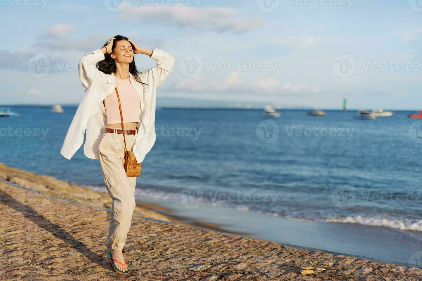 een vrouw gelukkig met haar ogen Gesloten in de zon wandelen Aan de strand met haar armen verspreiding uit tegen de oceaan met een glimlach, vliegend haar, gebruind huid, ontspanning, een reis naar de oceaan en vrijheid. foto
