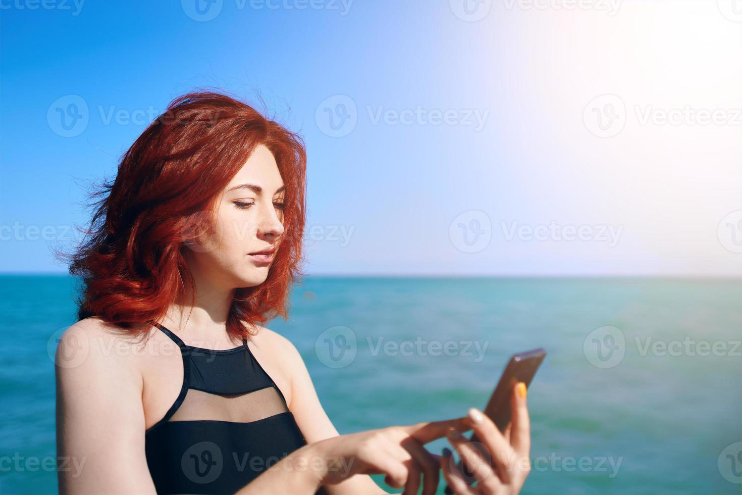 roodharige vrouw schrijft bericht op haar mobiele telefoon op de achtergrond van de zee-zonsondergang foto