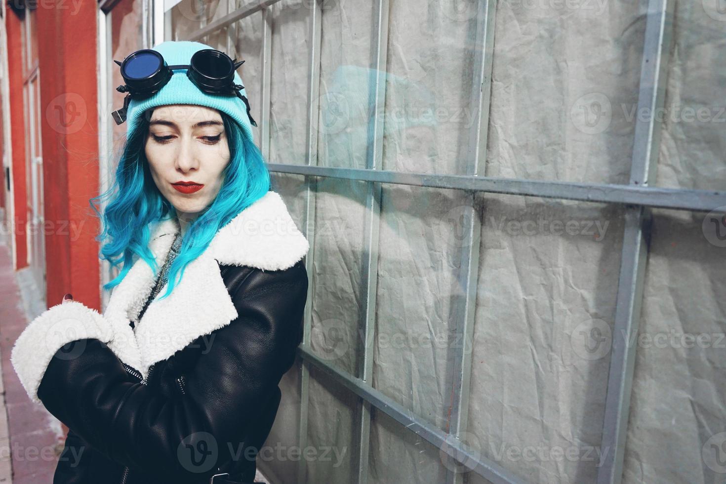 portret van een punk of gothic jonge vrouw met blauw gekleurd haar en het dragen van een zwarte steampunk bril en blauwe wollen muts in een stedelijke buitenstraat foto