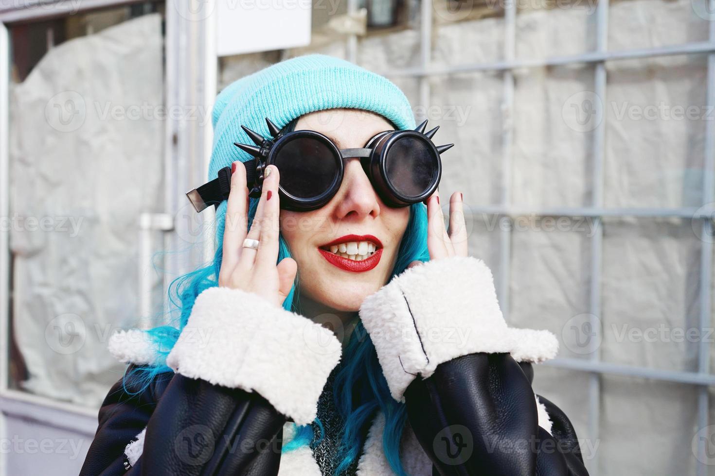 portret van een punk of gothic jonge vrouw glimlacht met blauw gekleurd haar en het dragen van een zwarte steampunk bril en blauwe wollen muts in een stedelijke buitenstraat foto