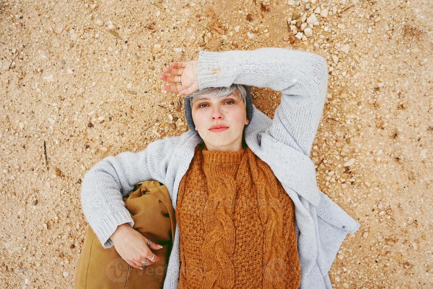 een jonge kaukasische avonturier die op zandgrond ligt naast een rugzak met een wollen trui en grijze wollen muts met oranje als hoofdkleur foto