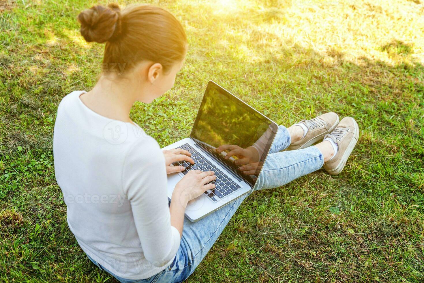 jonge vrouw zittend op groen gras gazon in stadspark werken op laptop pc-computer. freelance bedrijfsconcept foto