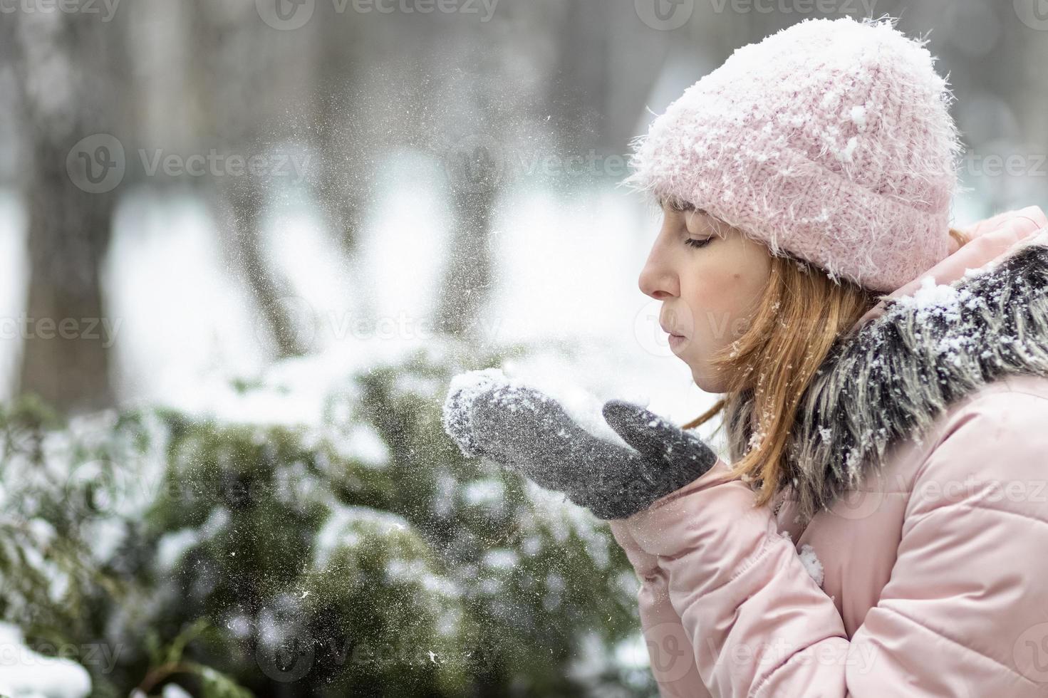 gelukkige vrouw op een besneeuwde winterdag in het park, gekleed in warme kleren, blaast de sneeuw van haar wanten foto