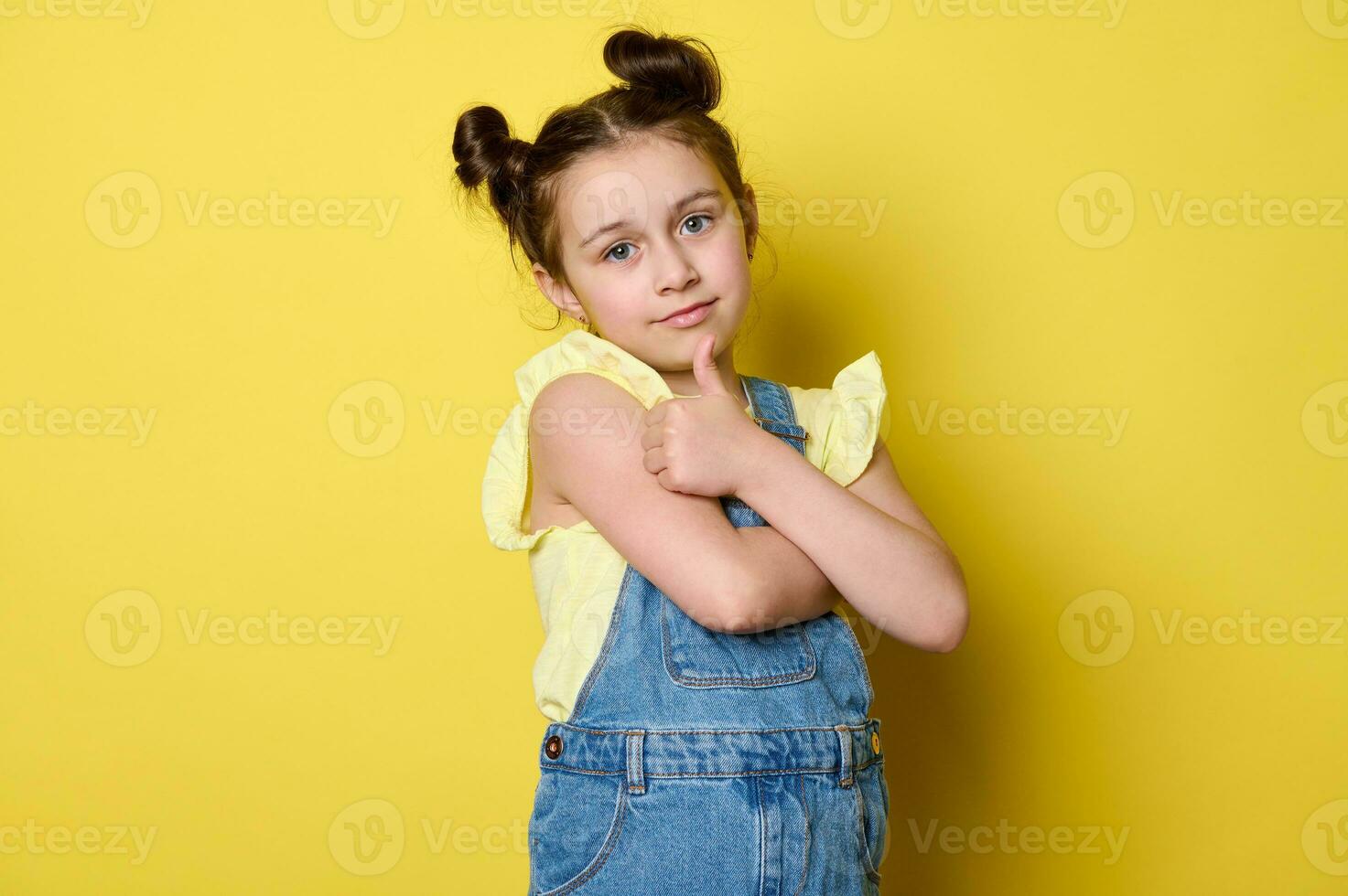 aanbiddelijk elegant kind meisje omarmen haarzelf en gebaren met duim omhoog, op zoek Vrolijk, geïsoleerd Aan geel achtergrond foto