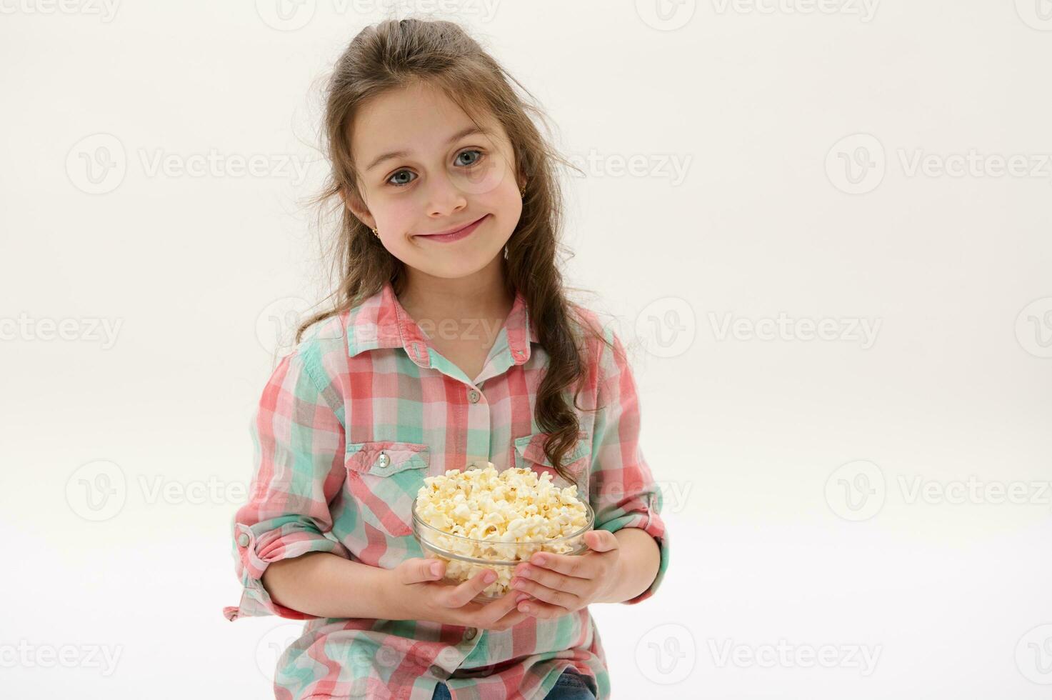 Kaukasisch weinig kind meisje glimlacht breed op zoek Bij camera, poseren met kom van zoet popcorn Aan wit studio achtergrond foto