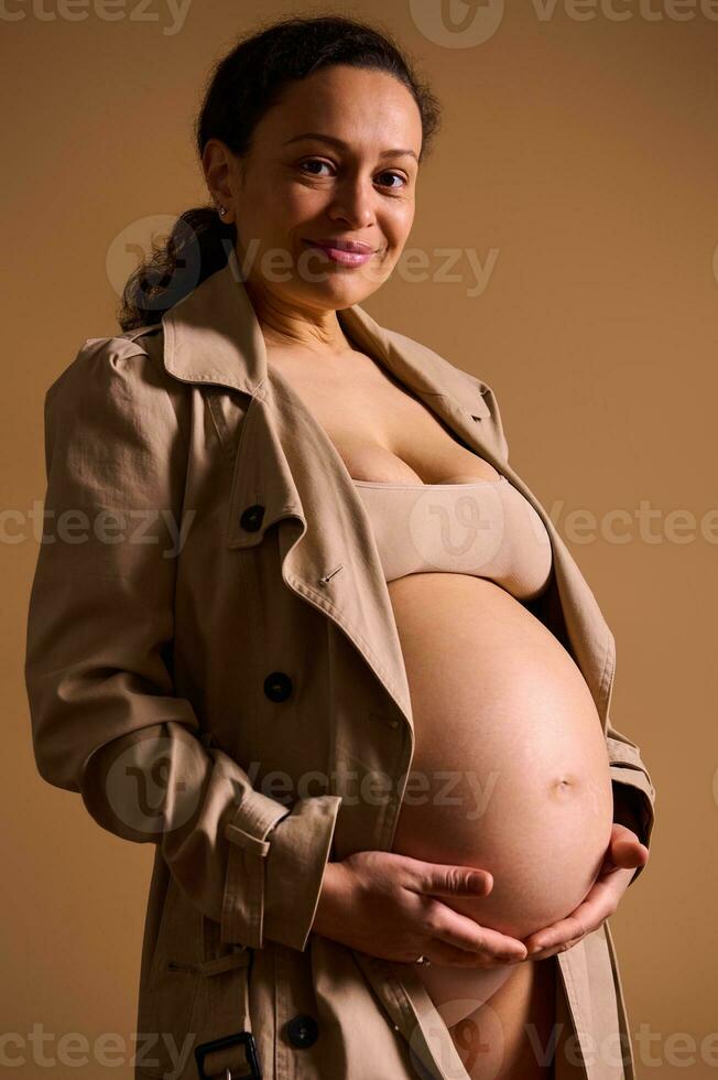 mooi zwanger vrouw in beige loopgraaf jas, voorzichtig strelen haar kaal buik, op zoek Bij camera, geïsoleerd Aan beige foto