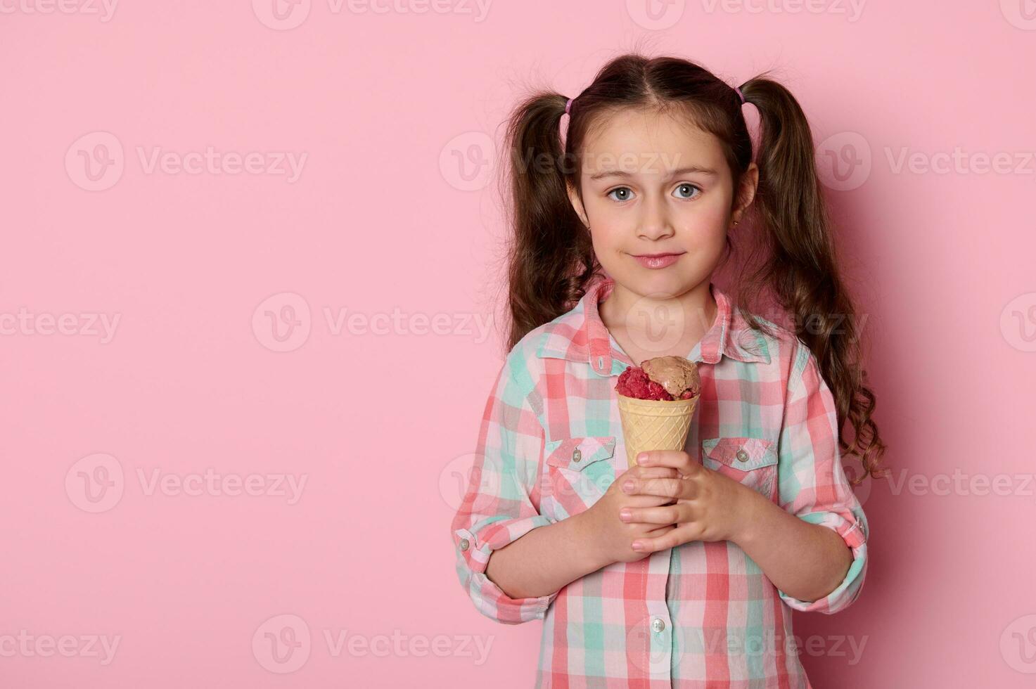 mooi weinig kind meisje met twee paardenstaarten, Holding een wafel ijs room, glimlachen op zoek Bij camera, geïsoleerd Aan roze foto