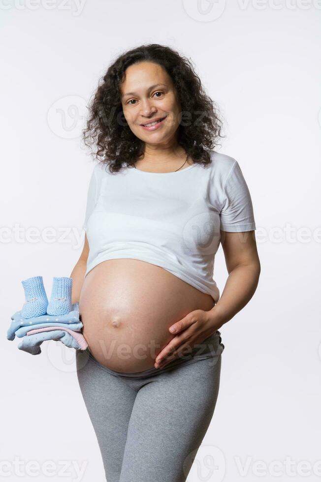 volwassen zwanger vrouw glimlacht op zoek Bij camera, strelen haar groot buik, Holding stapel van baby kleren Aan wit achtergrond foto