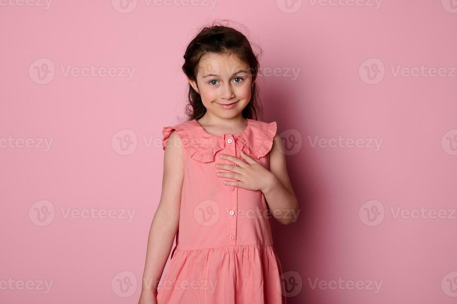 mooi verlegen weinig kind meisje in elegant roze jurk, glimlachen schattig op zoek Bij camera, geïsoleerd Aan roze achtergrond foto