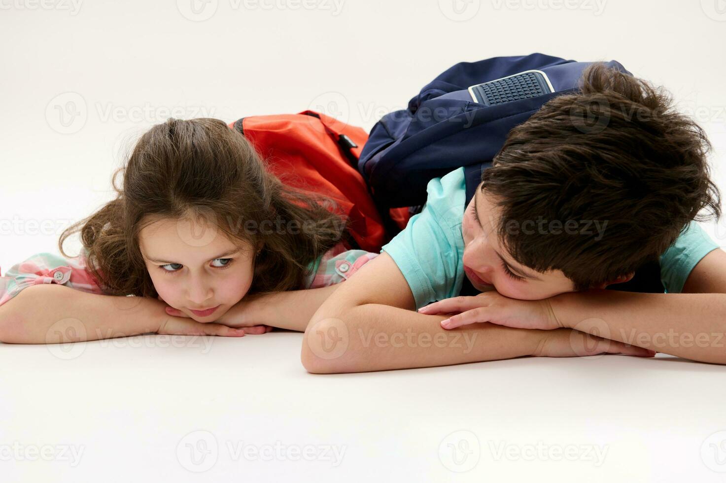 gelukkig weinig peuter- meisje en haar broer, tiener school- jongen met rugzakken aan het liegen Aan buik, geïsoleerd Aan wit achtergrond foto