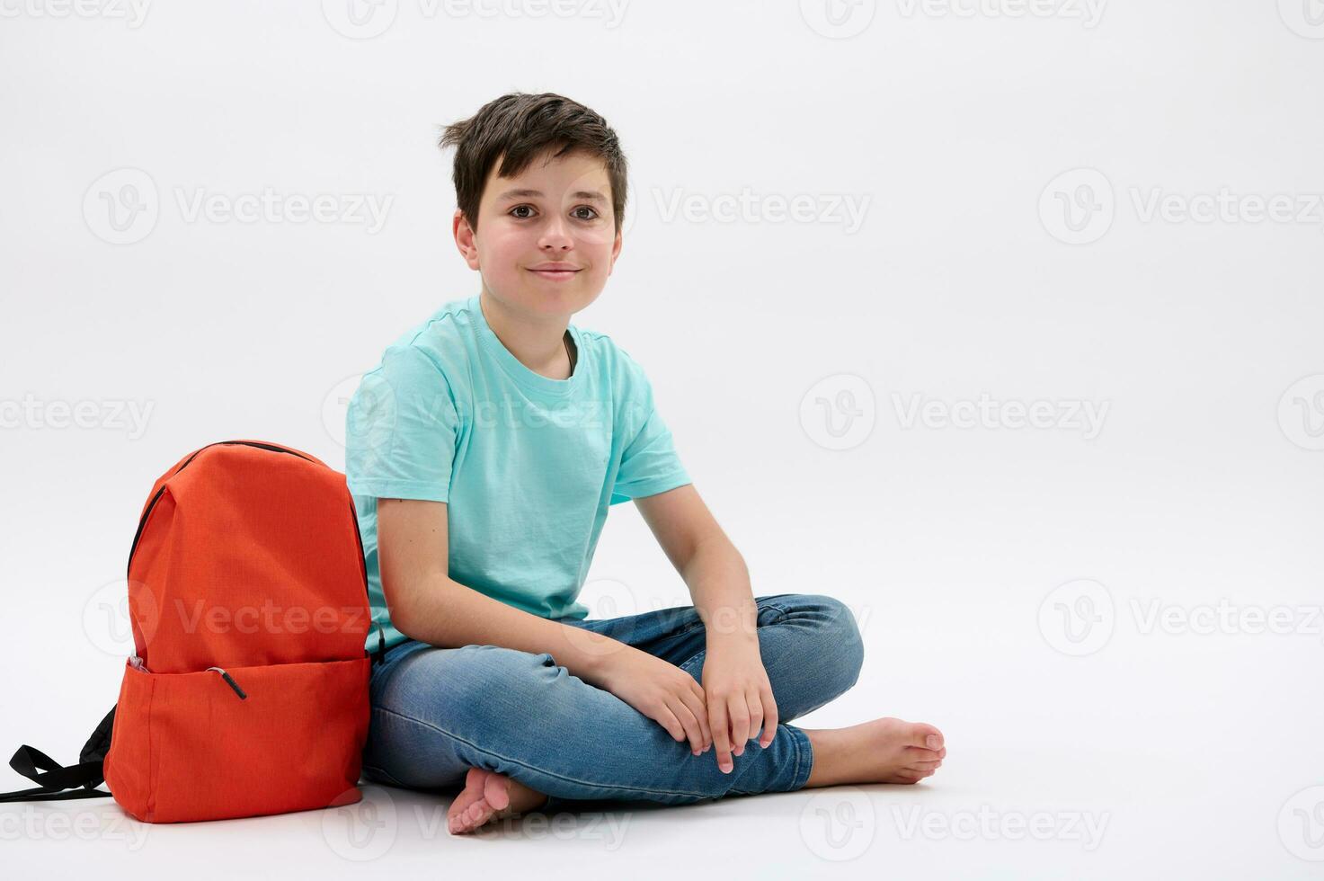 slim tiener jongen in blauw t-shirt en jeans met oranje rugzak, glimlacht op zoek Bij camera, geïsoleerd wit studio backdrop foto