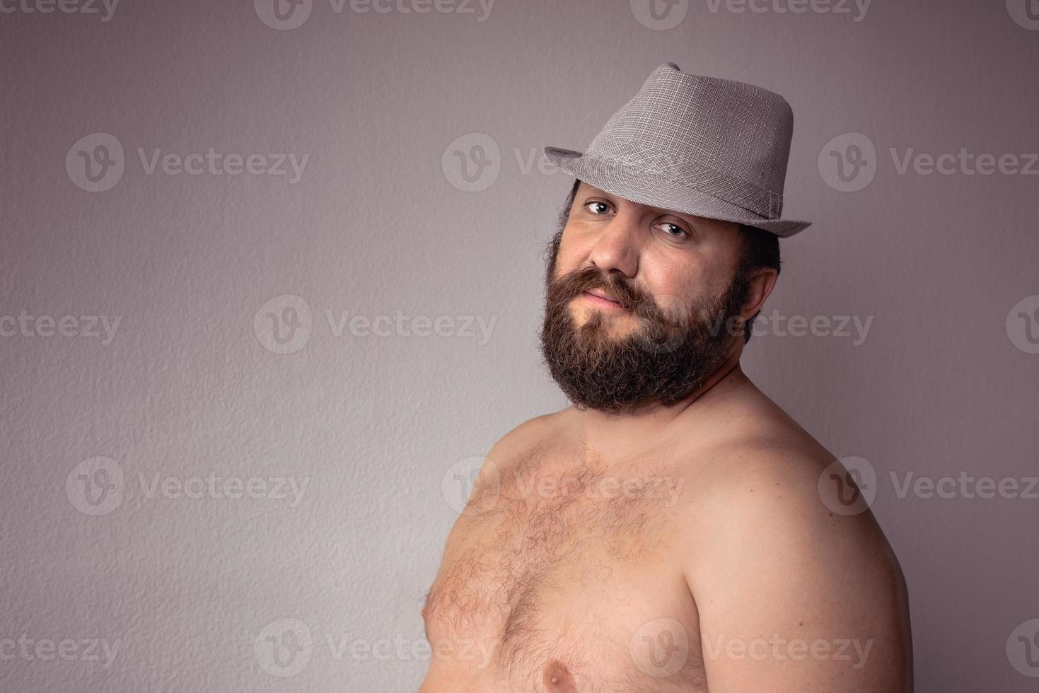 knappe halfnaakte bebaarde man met grijze hoed die tegen een grijze achtergrondmuur staat. foto