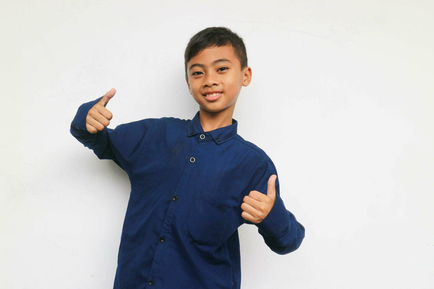 glimlachen zelfverzekerd jongen vervelend een blauw gewoontjes overhemd op zoek Bij de camera met tonen twee duim omhoog geïsoleerd Aan wit achtergrond foto