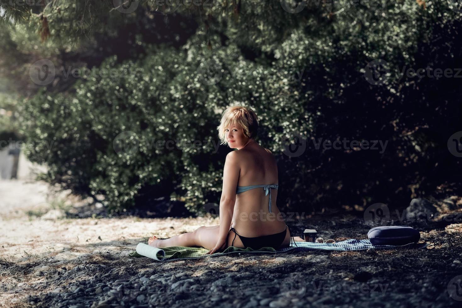 vrouw in bikini alleen genieten op het strand. blonde vrouw genieten van haar koffie in de schaduw van dennenboom op het wilde strand. foto