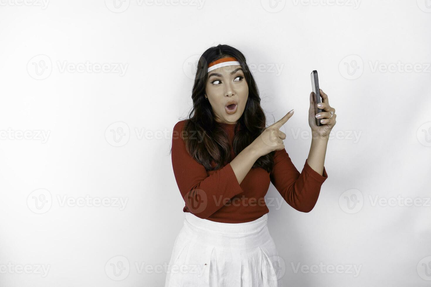 een portret van een geschokt Aziatisch vrouw vervelend hoofdband, Holding haar telefoon terwijl haar mond breed open, geïsoleerd door wit achtergrond. Indonesië's onafhankelijkheid dag concept foto