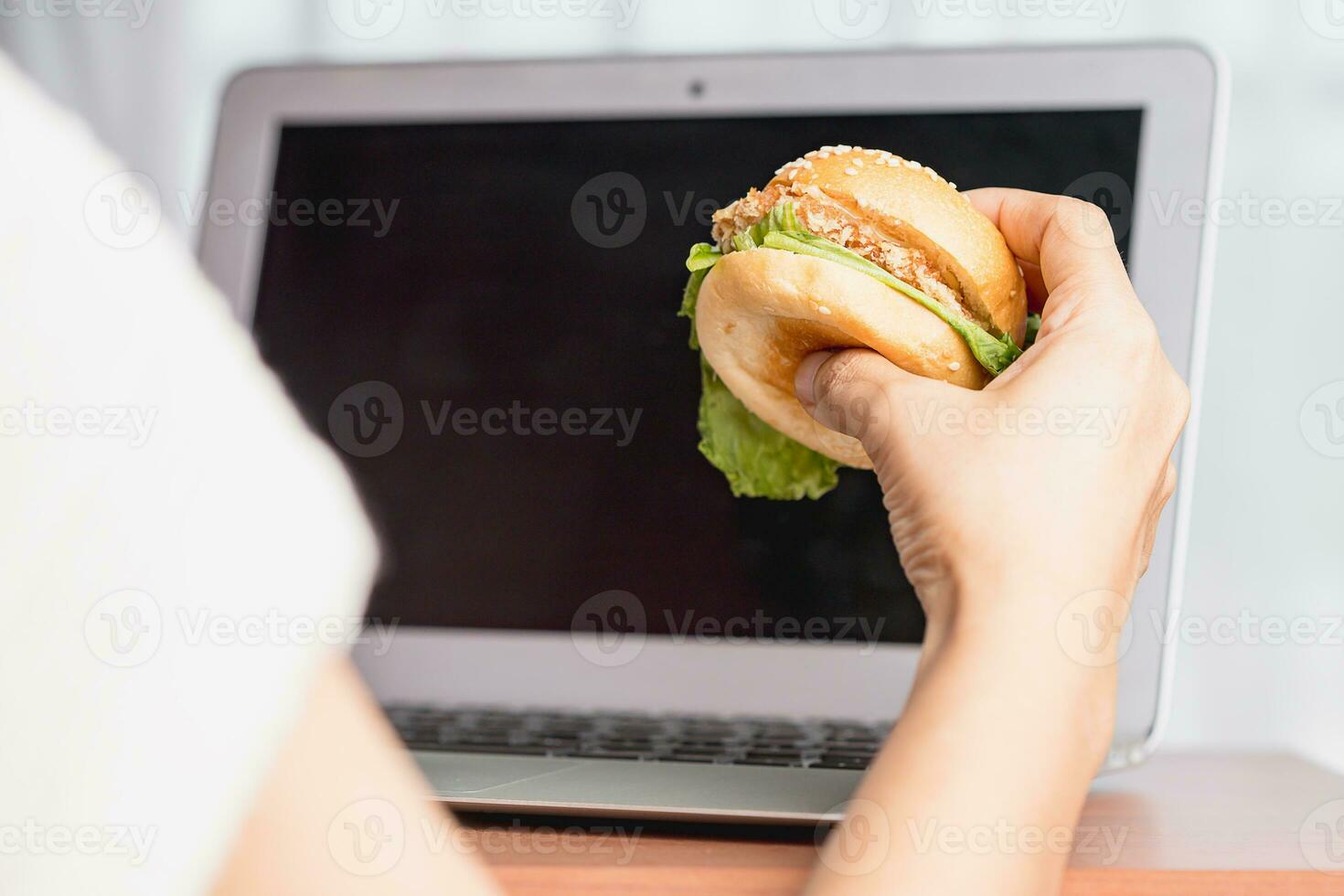 hand- Holding een heerlijk Hamburger en gebruik makend van laptop computer foto