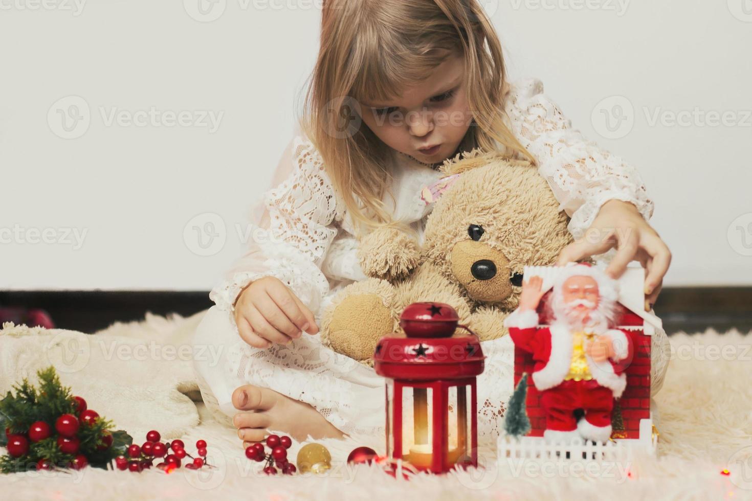 schattig klein blond meisje zittend op de vloer, spelend met haar teddybeer en kleine kerstman speelgoed foto