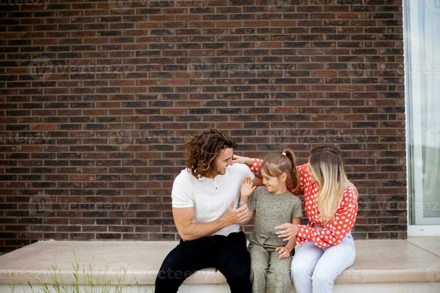 familie met een moeder, vader en dochter zittend buiten Aan de stappen van een voorkant veranda van een steen huis foto