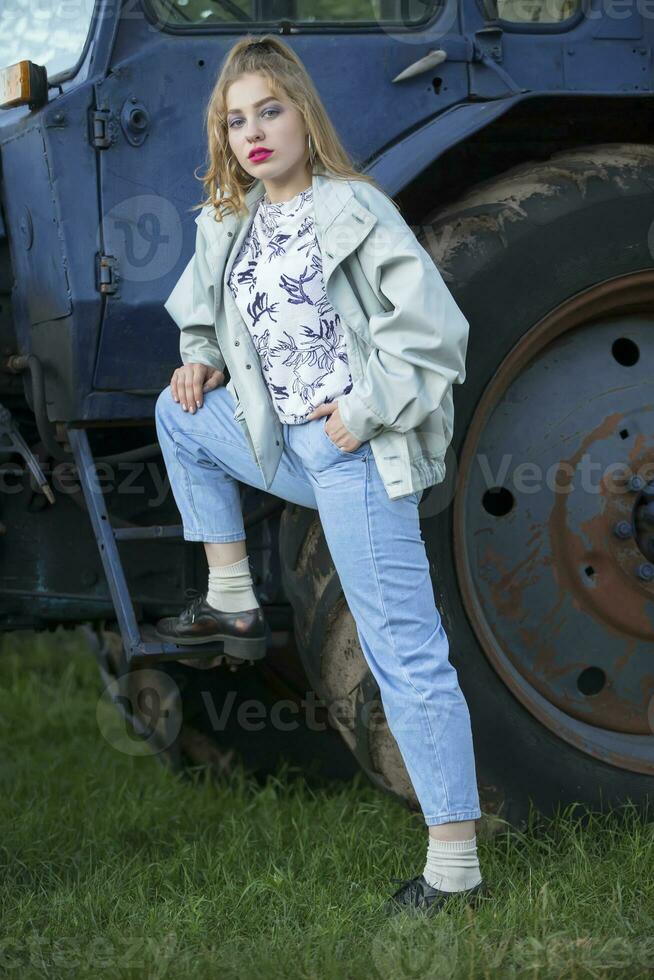 een mooi land meisje in de stijl van de 90s in helder kleren staat in de buurt een tractor. foto