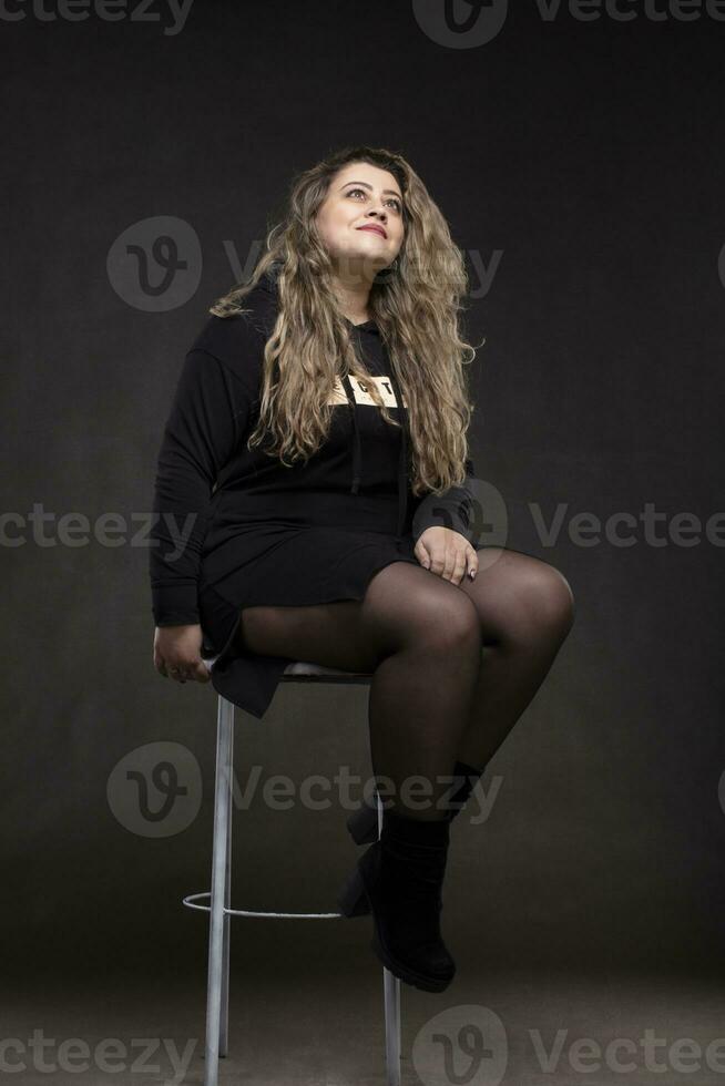 mooi dik vrouw met lang gekruld haar- in zwart kleren Aan een grijs achtergrond. plus grootte model- meisje. foto