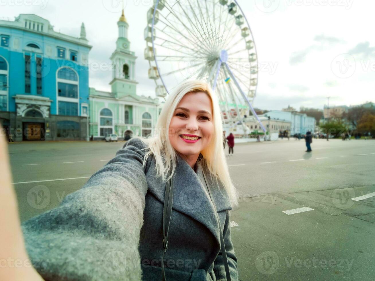 aantrekkelijk blond meisje in gewoontjes kleding, vervelend een jas, duurt selfie, op zoek in de camera. elegant meisje duurt selfie, achtergrond van de ferris wiel foto