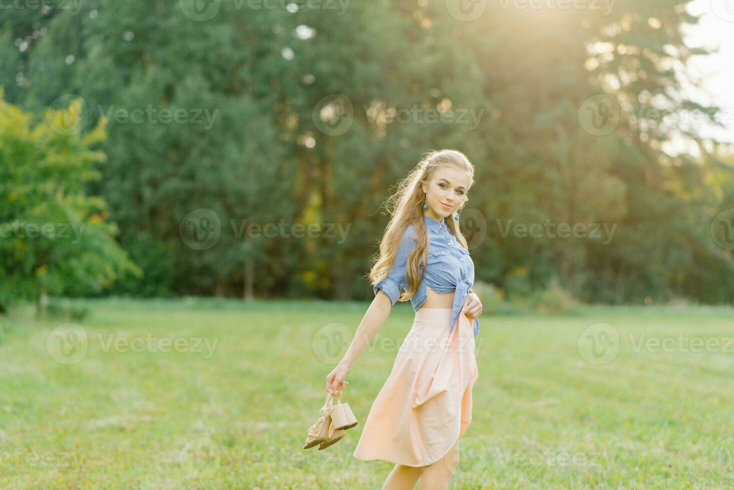 jong Kaukasisch vrouw of meisje in een zomer jurk en een denim overhemd is wandelen Aan de gras, Holding schoenen in haar handen, ontspannende buiten de stad Aan een weekend in zomer foto