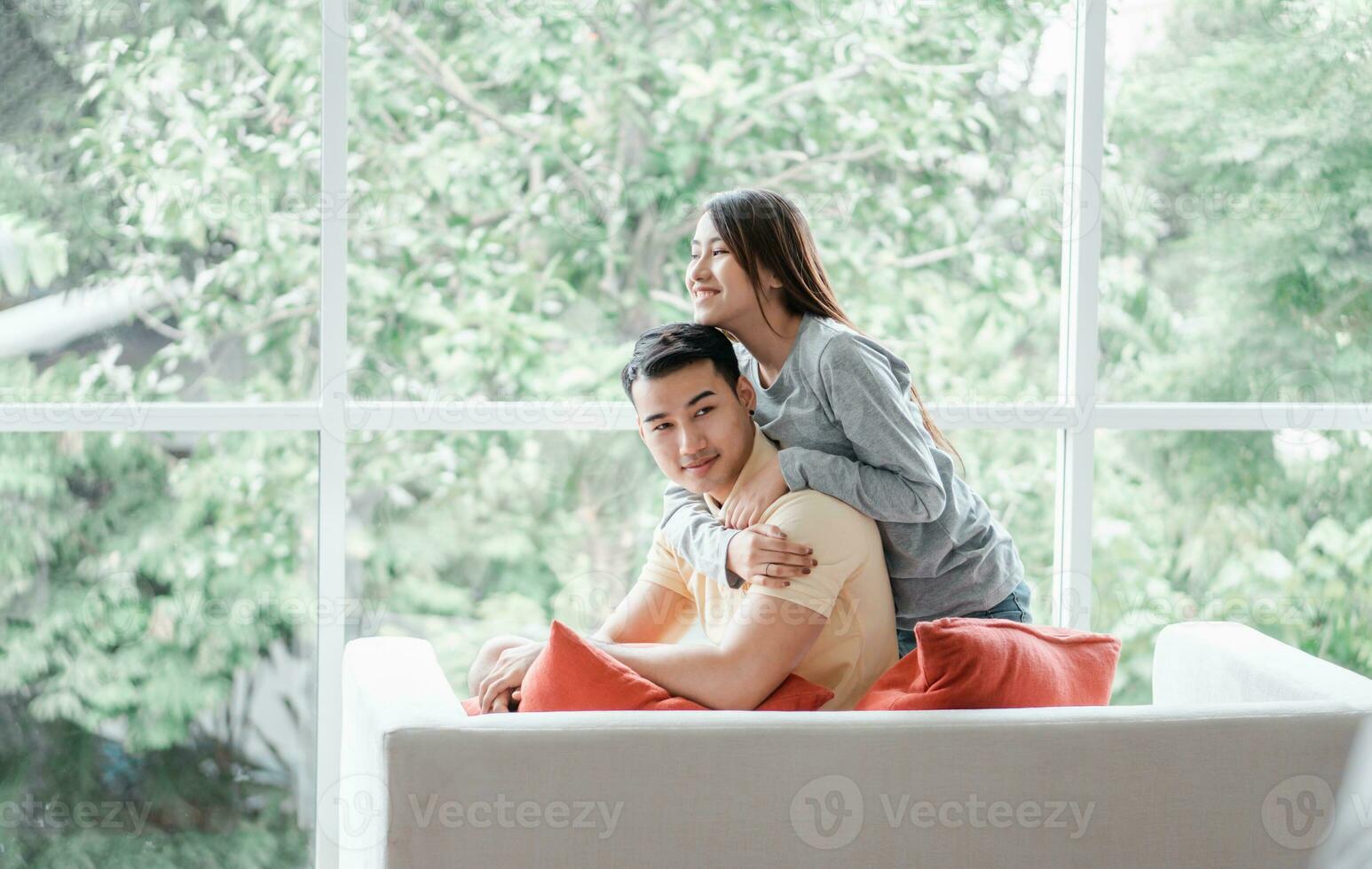 gelukkig paar zittend Aan de sofa en wezen een vrouw omarmen haar vriendje met liefde in de leven kamer en glimlach. concept van romantisch Aan Valentijn dag. voorstel en huwelijk foto