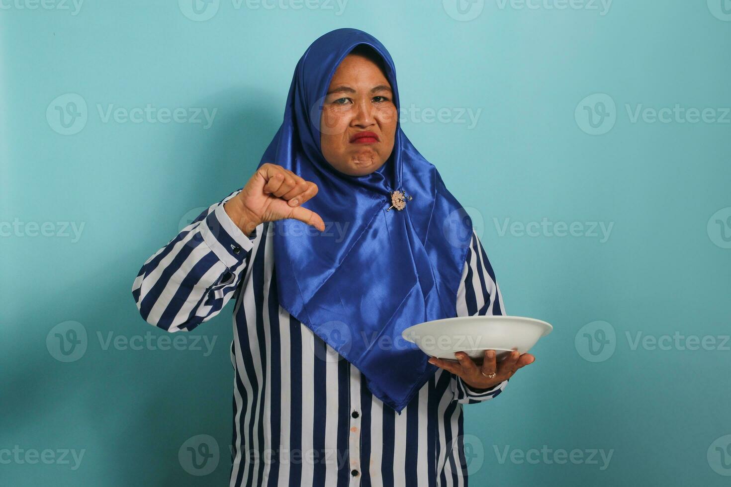 een boos middelbare leeftijd Aziatisch vrouw, vervelend een blauw hijab en gestreept shirt, wordt weergegeven een ongelukkig uitdrukking, shows duimen omlaag, en houdt een wit bord, geïsoleerd tegen een blauw achtergrond foto