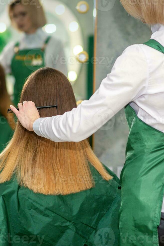 een vrouw kapper is kammen de lang bruin haar- van een jong vrouw Bij een salon. foto