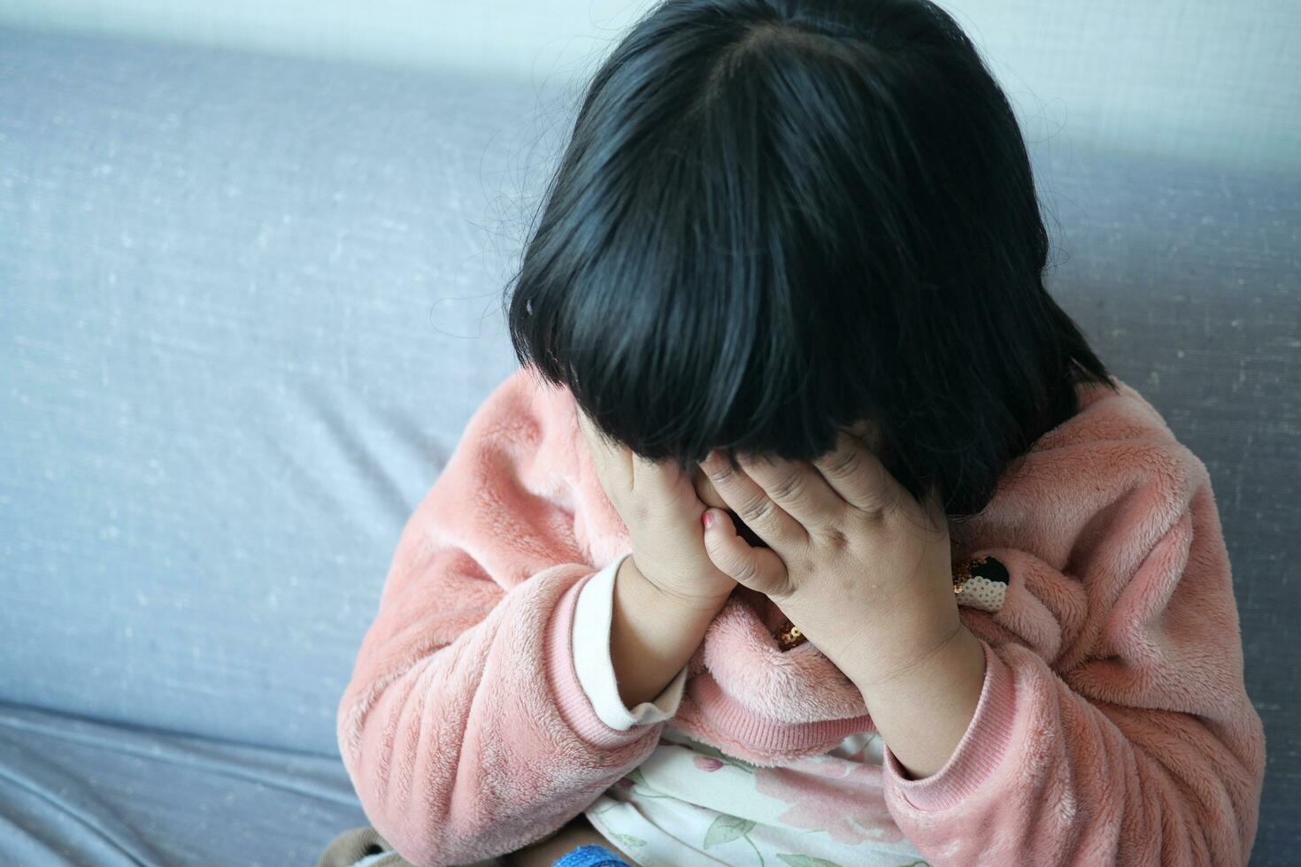 een van streek kind meisje Hoes haar gezicht met hand- zittend Aan sofa Bij huis foto