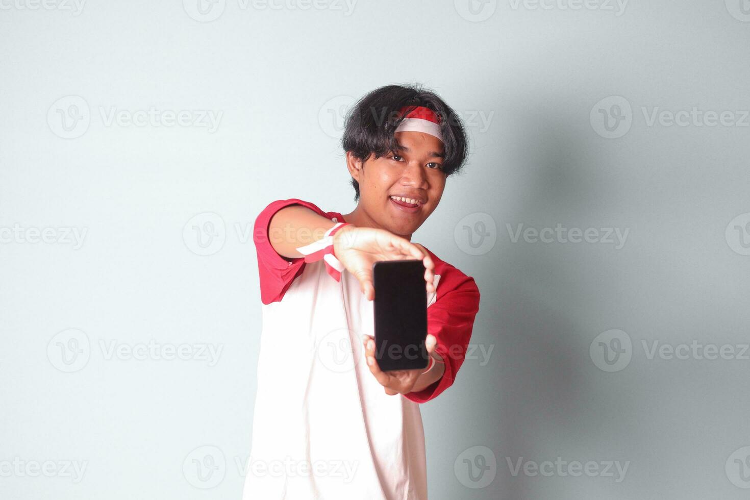 portret van aantrekkelijk Aziatisch Mens in t-shirt met rood en wit lint Aan hoofd, Holding en tonen blanco scherm van mobiel telefoon voor maquette. geïsoleerd beeld Aan grijs achtergrond foto