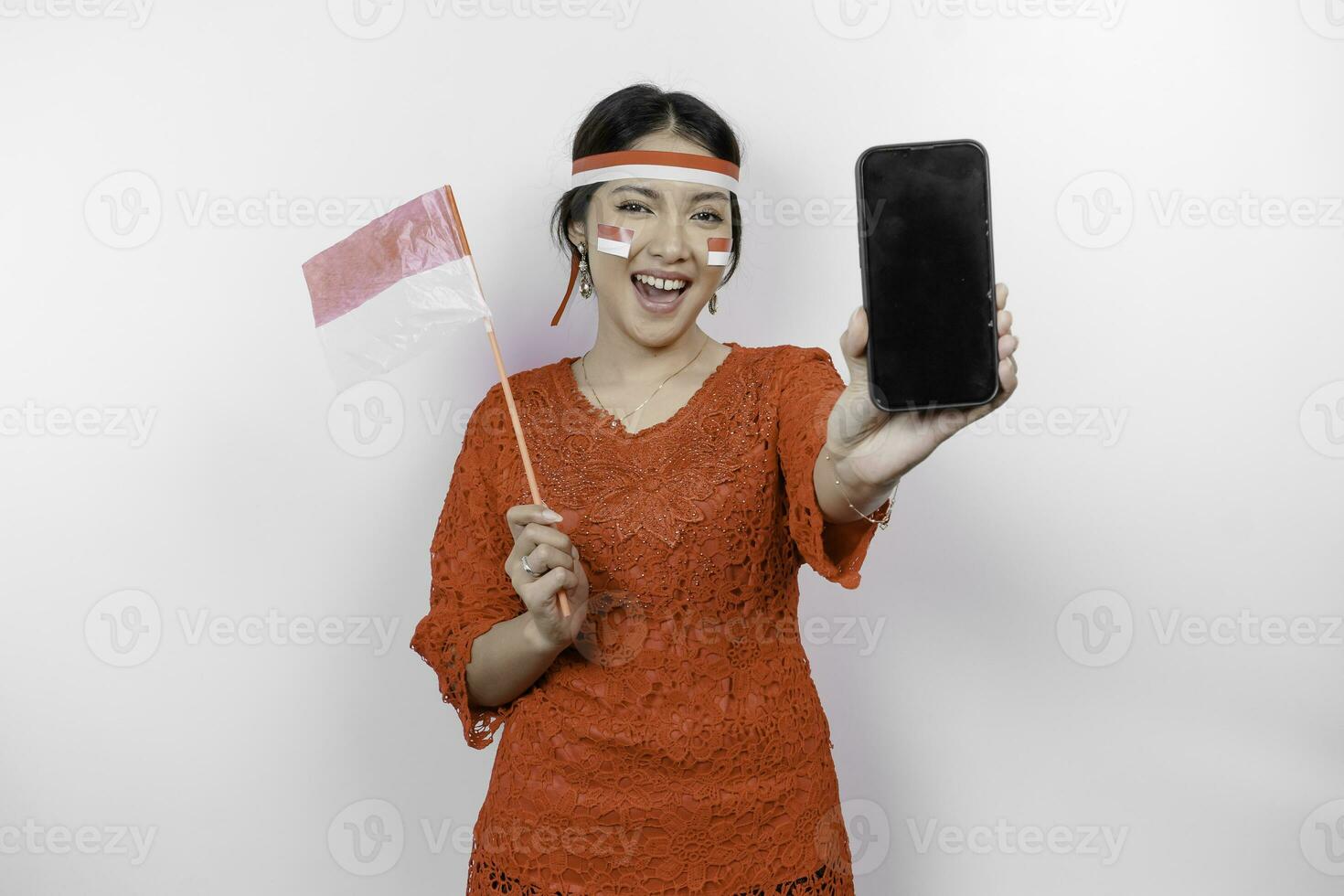 een portret van een glimlachen Aziatisch vrouw vervelend rood kebaya en hoofdband en tonen haar telefoon, geïsoleerd door wit achtergrond. Indonesië's onafhankelijkheid dag concept foto