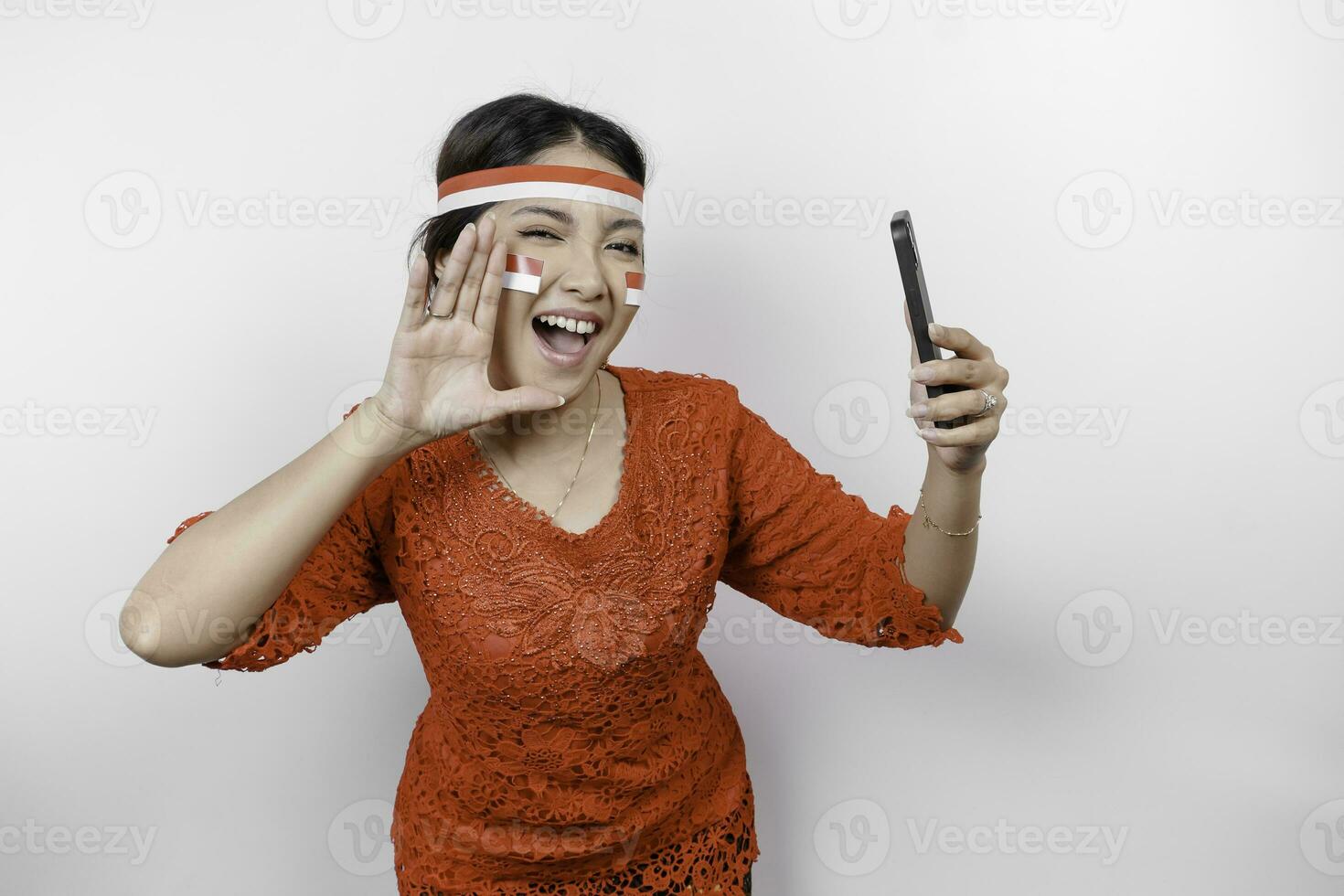 jong mooi vrouw vervelend een rood kebaya is Holding haar telefoon terwijl geschreeuw en schreeuwen luid met een hand- Aan haar mond. Indonesië's onafhankelijkheid dag concept. foto