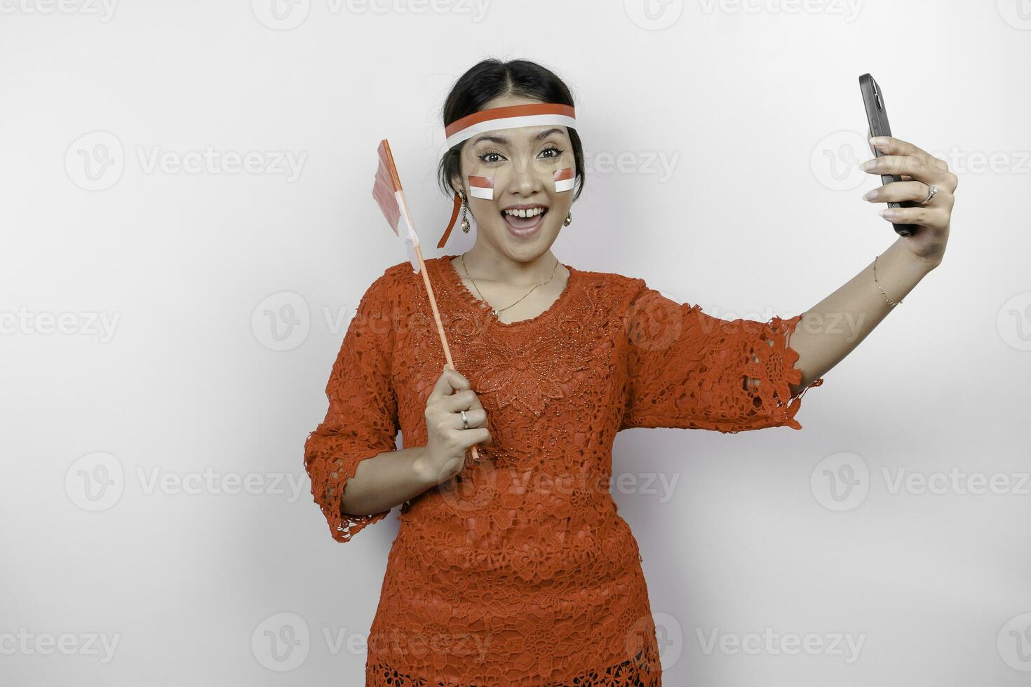 een gelukkig Aziatisch vrouw vervelend rood kebaya en hoofdband, Holding haar telefoon en Indonesië's vlag, geïsoleerd door wit achtergrond. Indonesië's onafhankelijkheid dag foto