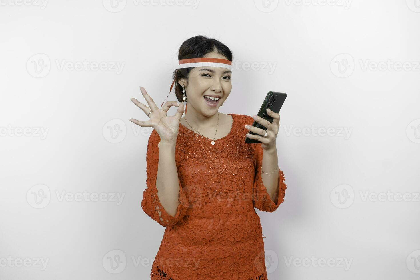een vrolijk Aziatisch vrouw vervelend rood kebaya en hoofdband, Holding haar telefoon terwijl gebaren OK teken met haar vingers, geïsoleerd door wit achtergrond. Indonesië's onafhankelijkheid dag concept foto