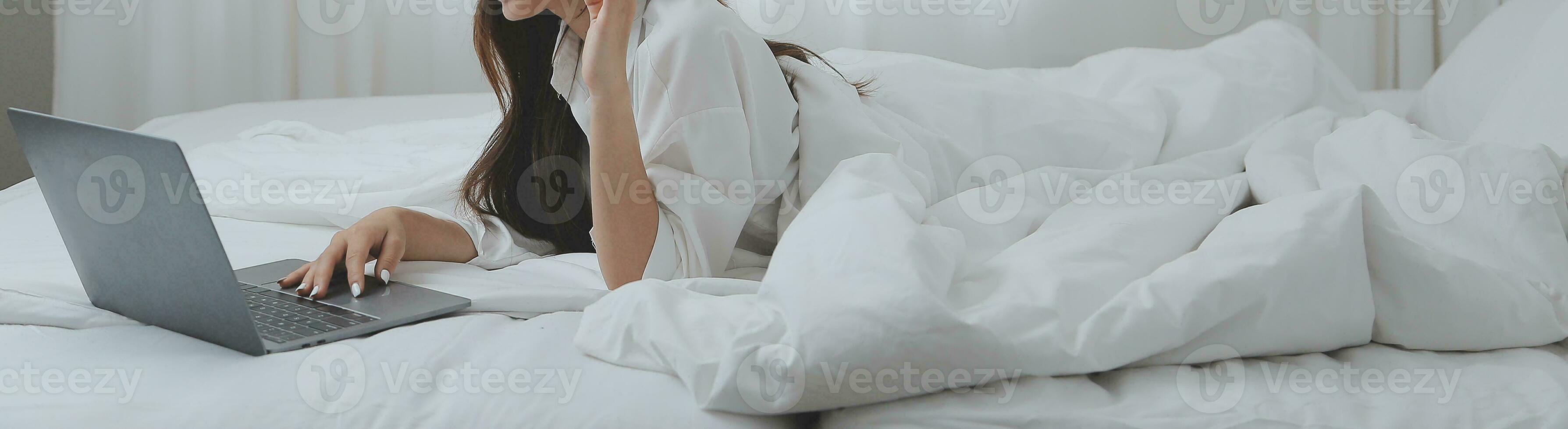 gelukkig jong vrouw in gewoontjes kleren glimlachen en op zoek Bij camera terwijl gebruik makend van laptop Aan bed in slaapkamer Bij huis foto