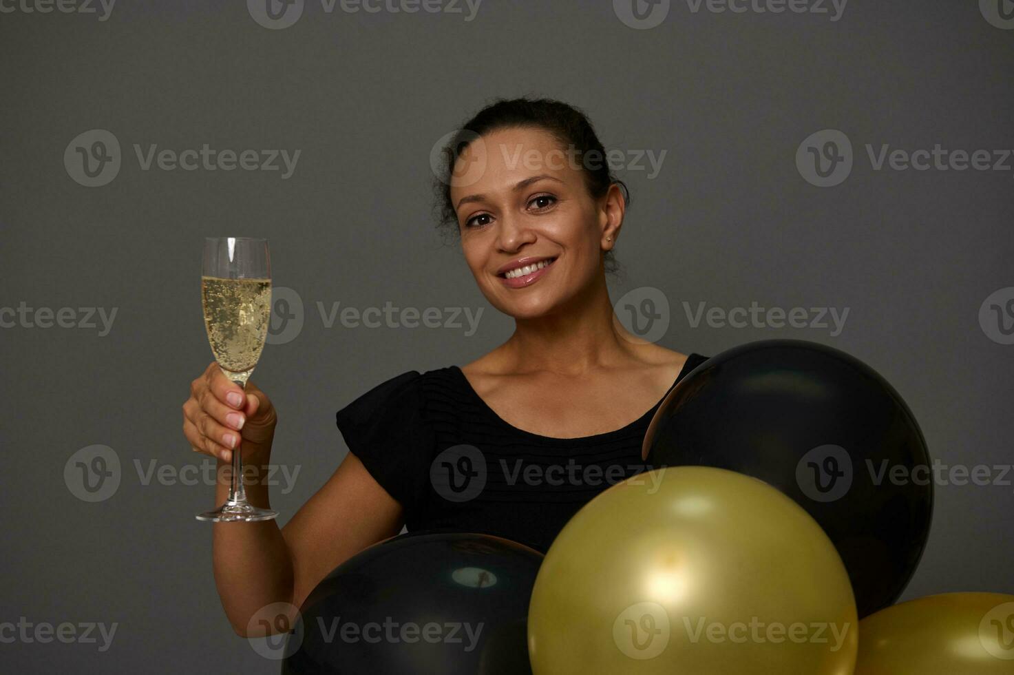 mooi glimlachen vrouw Holding een glas met sprankelend wijn poses met gouden en zwart opgeblazen lucht ballonnen tegen grijs muur achtergrond met kopiëren advertentie ruimte. concept van boodschappen doen Bij zwart vrijdag foto