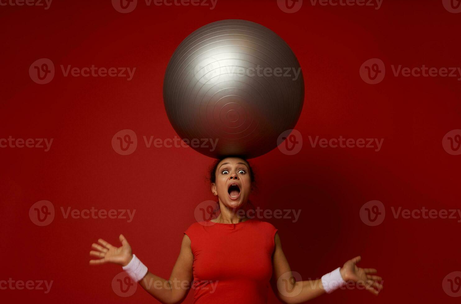 verbaasd jong sportief gemengd ras vrouw poses tegen rood muur achtergrond met reusachtig geschiktheid bal Aan haar hoofd foto