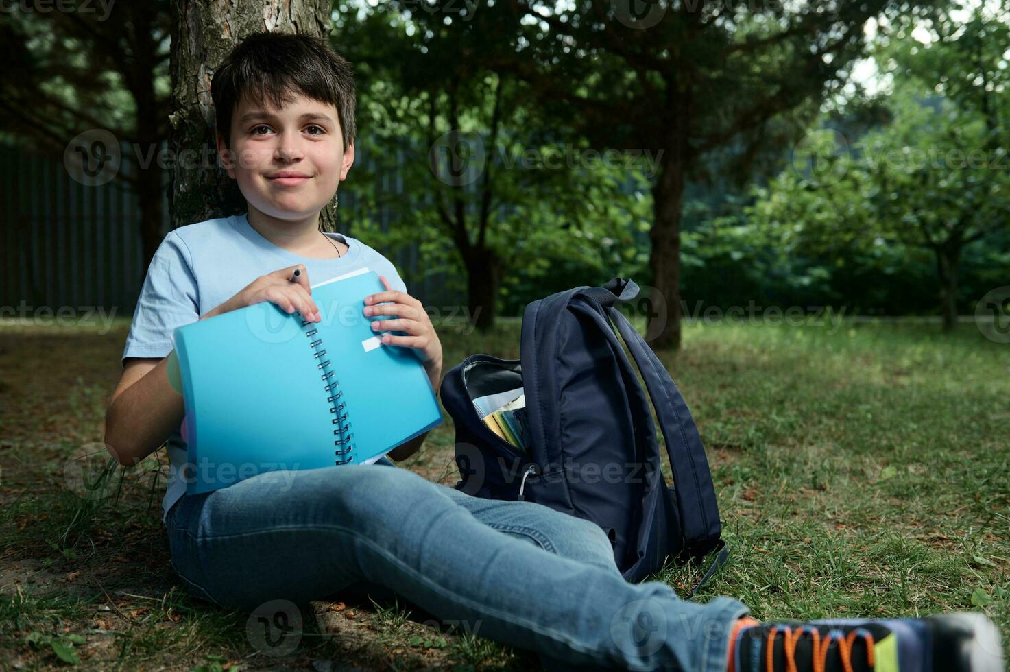 aanbiddelijk preteen schooljongen Holding schrijfboek, ontspannende in een park na klas, glimlacht schattig op zoek Bij camera. foto