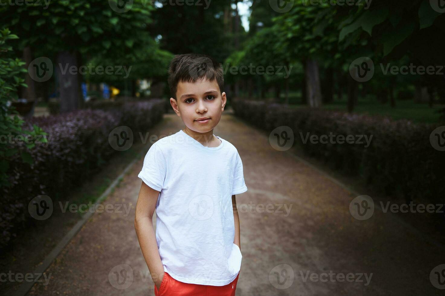 portret knap Kaukasisch school- leeftijd jongen 9 jaren oud, in wit t-shirt, op zoek Bij camera, staand Aan steeg van een park foto