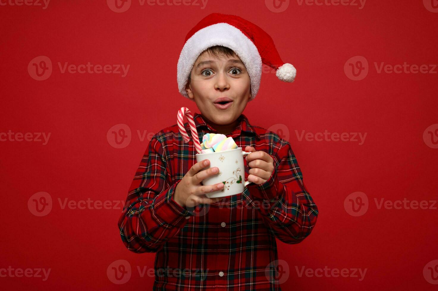 mooi versteld staan preteen jongen vervelend een de kerstman hoed poses tegen rood achtergrond met een kop van heet chocola drinken met marshmallows en gestreept zoet snoep riet. Kerstmis concept met kopiëren ruimte voor advertentie foto